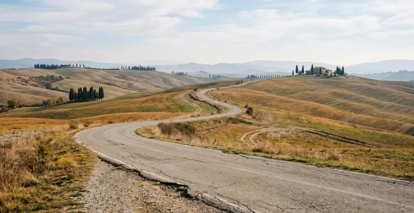 An empty winding country road through the rolling hills and cypress-lined landscape of Val d'Orcia, Tuscany.